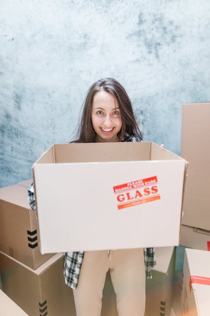 Two women in casual clothing are inside a room with a textured, light grey wall, surrounded by stacked cardboard moving boxes, some sealed with red and black tape. One woman with shoulder-length brown hair is holding a box, while the other woman, with curly black hair, is smiling and holding a large decorative vase with a plant arrangement. The floor is made of light wood, and paper packing peanuts are scattered on it. The scene appears to depict a home relocation process, with the women preparing for or engaged in packing and moving activities as part of a professional removals service, such as those provided by Removal Company Harringay. The setting suggests an organized and careful packing and loading process in a home or storage environment, relevant to house removals and furniture transport.