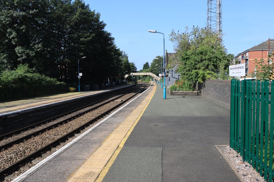 The image shows a quiet house removal and moving logistics scene at Crouch Hill Station in Harringay on a clear day. The platform is lined with a yellow tactile paving strip along the edge, adjacent to the railway tracks that run parallel and extend into the distance. To the right, a paved walkway is bordered by a green metal fence and a small garden with blooming plants and trees, some in partial shadow. A lamppost with a blue base stands along the platform, and further ahead, a station footbridge arches over the tracks. Residential buildings and a tall communication tower are visible beyond the station, providing context for urban house relocations. The environment appears well-lit with natural sunlight, emphasizing the cleanliness and orderliness of the station area, which may be relevant in the context of house removals and transportation planning coordinated by Removal Company Harringay.