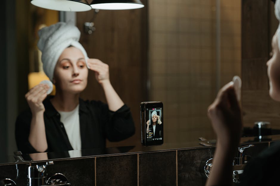 A woman with a white towel wrapped around her head is standing in front of a bathroom mirror, applying skincare or makeup, with her right hand holding a cosmetic sponge near her face. She is wearing a black top, and her reflection shows her looking intently at her face in the mirror. A smartphone is propped on the edge of the sink, capturing her image for a selfie or video. The bathroom has brown wooden panels on the walls, a white ceiling, and a dark-colored countertop below the mirror. The scene is illuminated by soft, warm lighting. This image is related to home relocation and personal grooming, which can be part of a moving or packing process, and it is provided by Removal Company Harringay, who specialise in removals and furniture transport services.