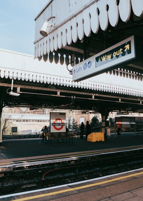 The image shows a quiet house removal and moving logistics scene at Crouch Hill Station in Harringay on a clear day. The platform is lined with a yellow tactile paving strip along the edge, adjacent to the railway tracks that run parallel and extend into the distance. To the right, a paved walkway is bordered by a green metal fence and a small garden with blooming plants and trees, some in partial shadow. A lamppost with a blue base stands along the platform, and further ahead, a station footbridge arches over the tracks. Residential buildings and a tall communication tower are visible beyond the station, providing context for urban house relocations. The environment appears well-lit with natural sunlight, emphasizing the cleanliness and orderliness of the station area, which may be relevant in the context of house removals and transportation planning coordinated by Removal Company Harringay.