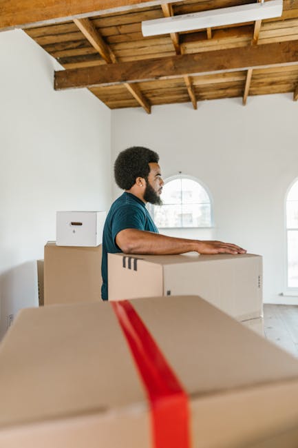 A man with dark hair and a beard, wearing a blue t-shirt, stands indoors in a room with white walls, wooden ceiling beams, and large arched windows. He is facing to the right, resting his hands on top