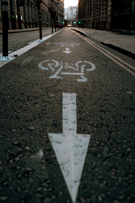 Urban street scene showing a designated bicycle lane with a white bicycle symbol and a downward pointing arrow painted on the asphalt. The lane is separated from the main road by black metal posts. In