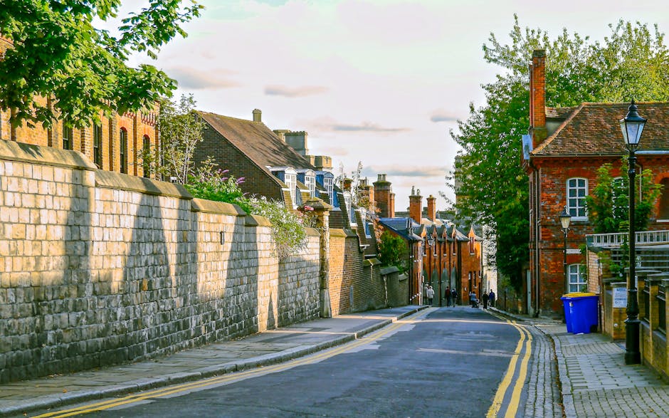 Urban street scene showing a designated bicycle lane with a white bicycle symbol and a downward pointing arrow painted on the asphalt. The lane is separated from the main road by black metal posts. In