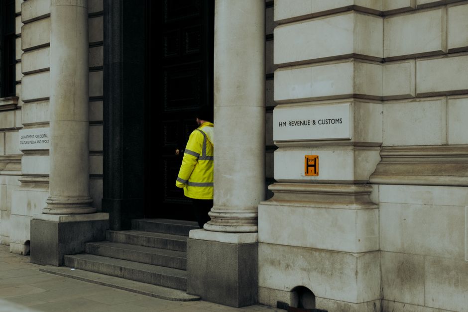 A personnel in a high-visibility yellow jacket with dark trousers is standing on stone steps at the entrance of a building with classical architectural features, including large stone columns and intricate stonework. The person appears to be waiting or observing, with their back turned toward the camera. The building’s facade is made of light-colored stone, with a black door in the center. To the right of the entrance, there is a white sign with black text reading 'HM REVENUE & CUSTOMS' attached to the wall, and a smaller orange label beneath it. The scene suggests a formal, government-related location, which could relate to the logistics or documentation involved in home relocation or moving services. Inside, the context could involve packing, moving, or transport procedures, commonly associated with house removals, supported by the professional services offered by Removal Company Harringay.