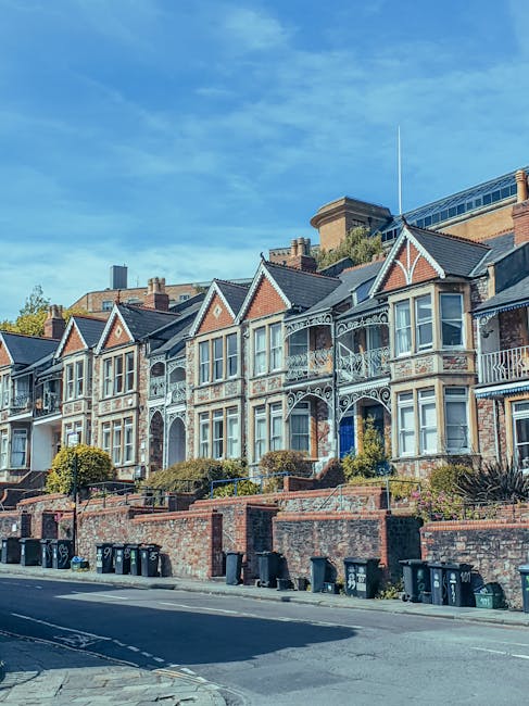 Image showing a row of Victorian-style terraced houses with ornate balconies and bay windows, situated on a residential street with brick front gardens and low brick walls. The houses are built with red bricks and feature gabled roofs with slate tiles. In front of the houses, there is a paved pavement with several black wheelie bins positioned along the curbside. The scene is captured during daylight under a partly cloudy sky, illustrating an urban neighbourhood environment typical of a house removal or home relocation setting. This image aligns with the context of local moving services offered by Removal Company Harringay, highlighting the transport and logistics involved in carrying out house removals in the area.