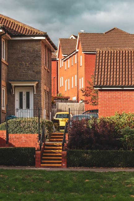 The image shows a residential area with a brick-built house on the left side, featuring a white front door and a small garden with green shrubs. A set of wooden steps with black metal railings leads up from the pavement to the house entrance, partially obscured by greenery. Behind the steps, a yellow car is parked on a driveway, with additional vehicles visible further back. In the background, there are several multi-storey houses with red brick walls and sloped, tiled roofs under a partly cloudy sky. The scene is well-lit, with natural daylight highlighting the details of the buildings, vehicles, and garden. The setting suggests a typical suburban neighbourhood suitable for house removals or furniture transport, with [COMPANY_NAME] likely involved in coordinating moving and packing services in this area, aligning with the context of house removals, stairs access, and logistics solutions described in the page title.