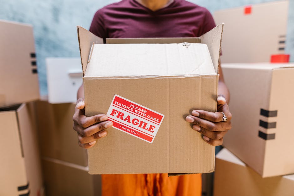 A person wearing a maroon shirt holding an open cardboard box with a red and white 'FRAGILE' sticker on the front, surrounded by other packed boxes in a moving or storage environment. The box is made of brown corrugated cardboard, partially filled with packing materials, and is positioned inside a room with additional cardboard boxes stacked nearby. In the background, there are more boxes with black tape seals, indicating a packing and moving process. The scene suggests careful handling and preparation for a home relocation, consistent with professional removals services from Removal Company Harringay, supporting efficient furniture transport and packing during house moves.