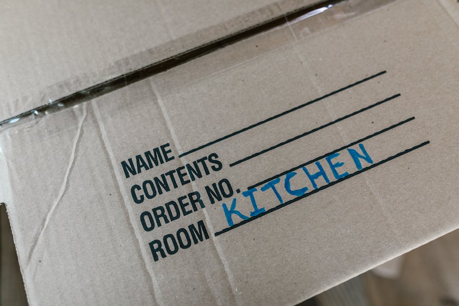Close-up of a cardboard moving box with printed labels indicating sections for name, contents, order number, and room, with 'KITCHEN' handwritten in blue marker on the 'ROOM' label. The box appears to be part of home packing and moving preparations, likely inside a property or on a moving trolley, with other boxes and packing materials possibly visible nearby. This scene illustrates the packing process involved in home relocation, as handled by Removal Company Harringay, supporting efficient furniture transport and interior moving logistics within the context of local house removals and packing service preparations.