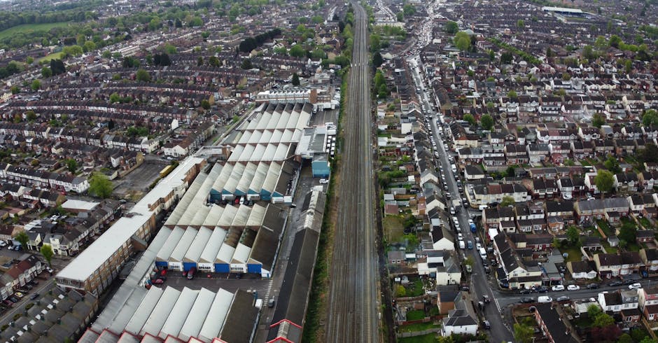 An aerial view of a residential neighbourhood with rows of terraced houses, detached homes, and green trees lining the streets on either side of a railway track running vertically through the centre. To the left of the railway, there is a large industrial warehouse with a series of interconnected, pitched-roof structures made of metal and concrete, adjacent to a loading area with visible trucks and cargo. Inside the warehouse area, forklifts and workers are seen handling cardboard boxes, furniture, and packaging materials, while nearby trolleys and straps are used for loading and securing items. The surrounding streets feature parked cars and driveways, with some doors open for access. The environment is well-lit with natural daylight, suggesting daytime hours, and the scene depicts a house removal and furniture transport process coordinated by Removal Company Harringay, involving packing, loading, and logistics within a busy urban setting.