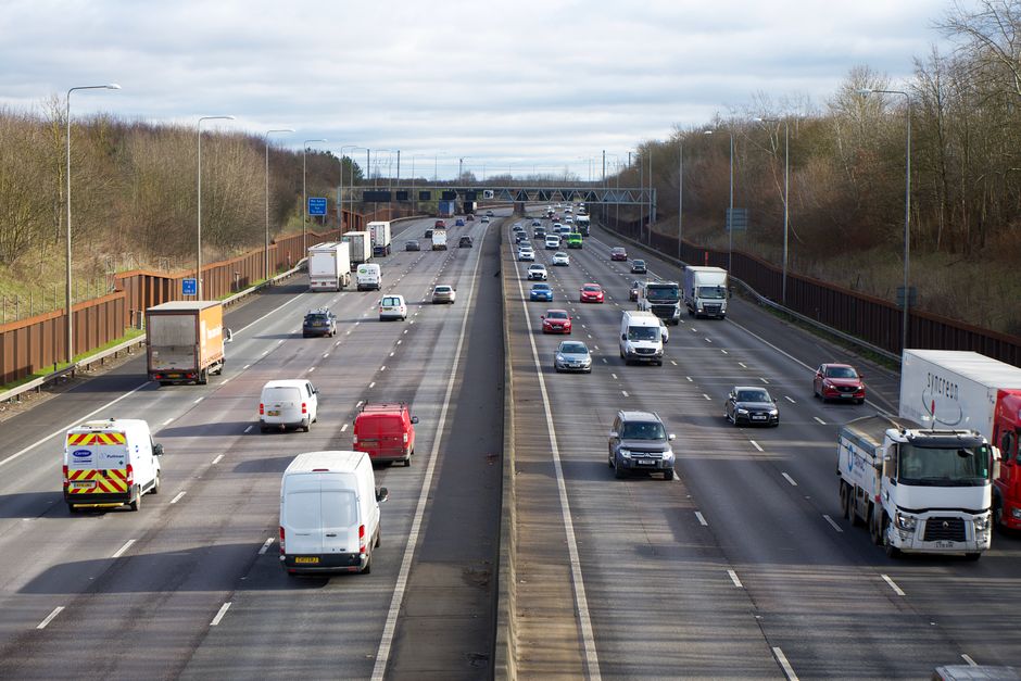 A view of a multi-lane motorway with various vehicles including trucks, vans, and passenger cars traveling in both directions. The road is divided by a central barrier, and overhead gantries and signage are visible in the distance. On the left side, the shoulder features a white van with a yellow and red striped warning pattern, and another white vehicle. The scene is outdoors during daytime with overcast skies, and surrounding areas include trees and streetlights. This image illustrates typical transport activity related to home relocation logistics, reflecting furniture transport and packing and moving processes managed by removals companies like Removal Company Harringay.