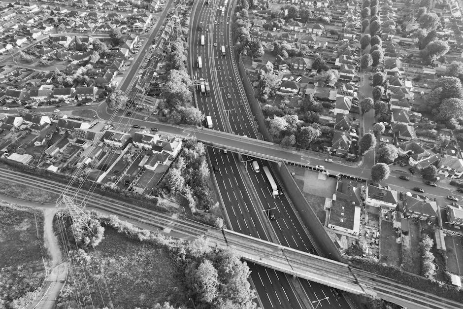 An aerial view of a residential neighbourhood with rows of terraced houses, detached homes, and green trees lining the streets on either side of a railway track running vertically through the centre. To the left of the railway, there is a large industrial warehouse with a series of interconnected, pitched-roof structures made of metal and concrete, adjacent to a loading area with visible trucks and cargo. Inside the warehouse area, forklifts and workers are seen handling cardboard boxes, furniture, and packaging materials, while nearby trolleys and straps are used for loading and securing items. The surrounding streets feature parked cars and driveways, with some doors open for access. The environment is well-lit with natural daylight, suggesting daytime hours, and the scene depicts a house removal and furniture transport process coordinated by Removal Company Harringay, involving packing, loading, and logistics within a busy urban setting.