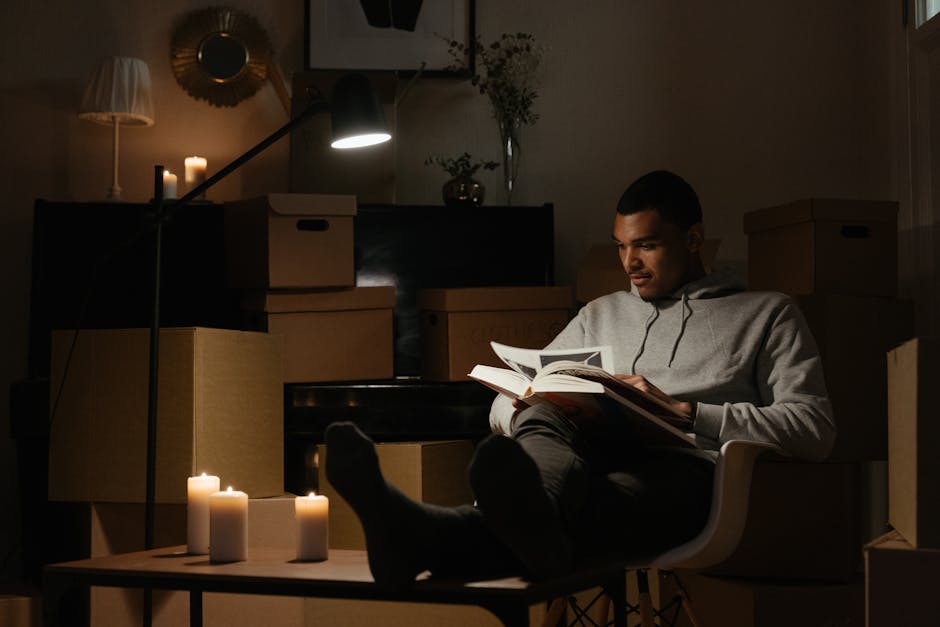 Two individuals, one wearing a grey long-sleeved shirt and the other in a white t-shirt and beige trousers, are seated on a wooden floor inside a property, engaged in packing or organizing household i