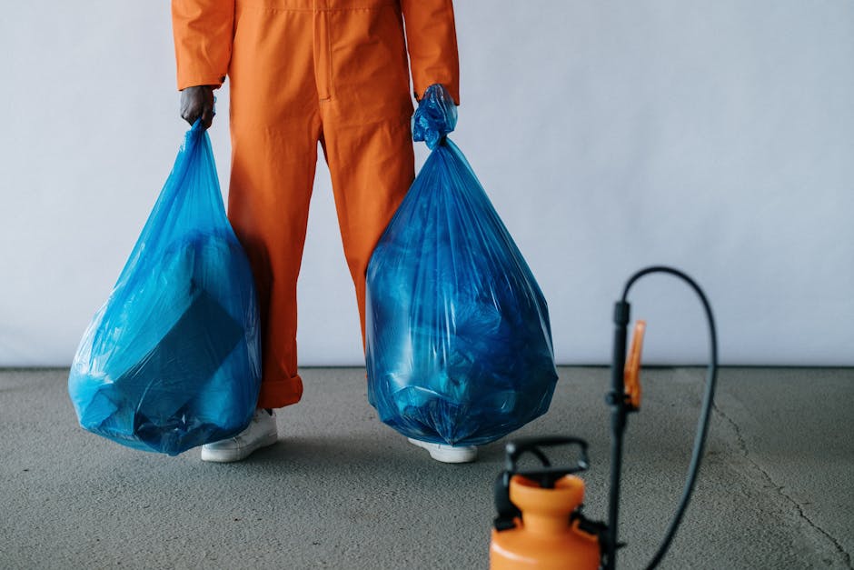 Close-up of a person's arm wearing a blue glove, reaching down to pick up a clear plastic bottle from a grassy outdoor area. In the background, a yellow plastic bag filled with trash and other waste i